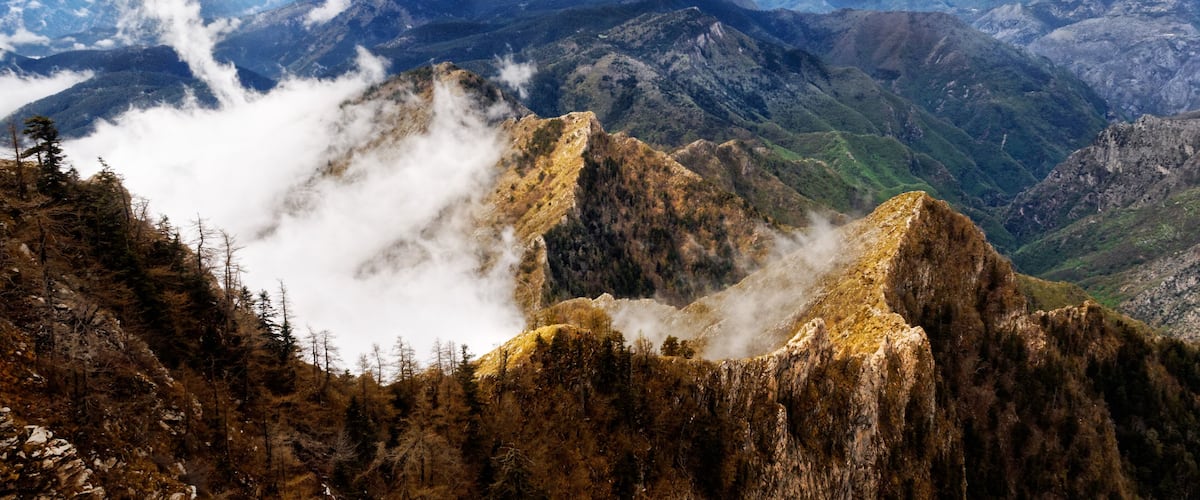 A mountainous landscape with rugged peaks, dense forests, and mist-covered valleys.