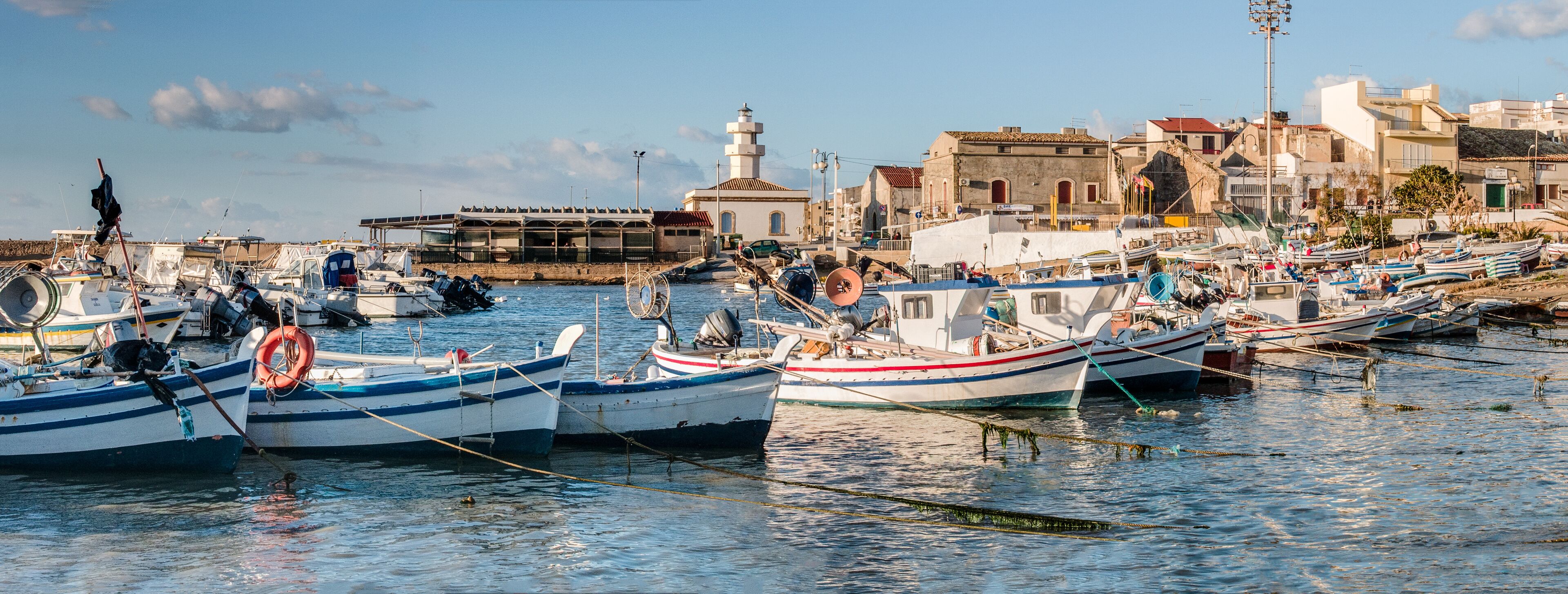 A harbor with numerous small boats docked, a lighthouse, and buildings in the background.