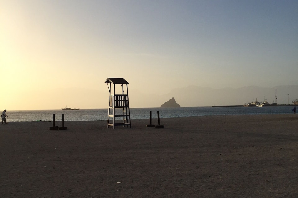 A beach with a lifeguard tower, a distant mountain, and a few boats in the water.