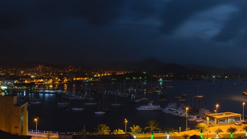 A nighttime marina with numerous boats docked, a cityscape in the background, and a mountain range.