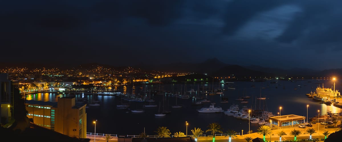 A nighttime marina with numerous boats docked, a cityscape in the background, and a mountain range.