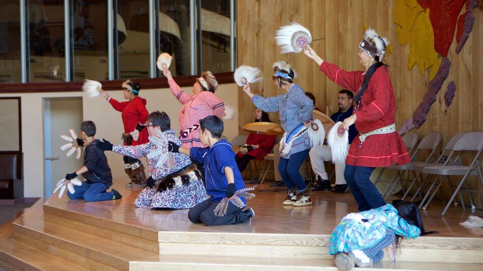 A group of people performing a traditional dance on a wooden stage with chairs and a wooden wall in the background.