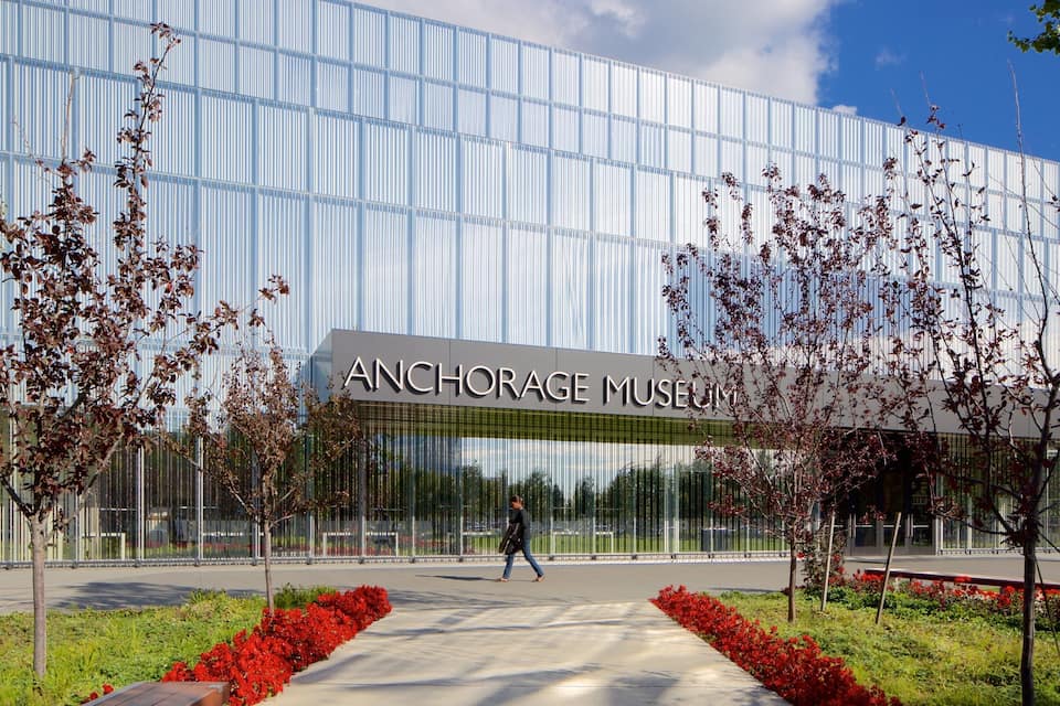 L’extérieur du musée d’Anchorage se distingue par sa façade moderne en verre, un chemin piétonnier et des plantes à fleurs rouges.