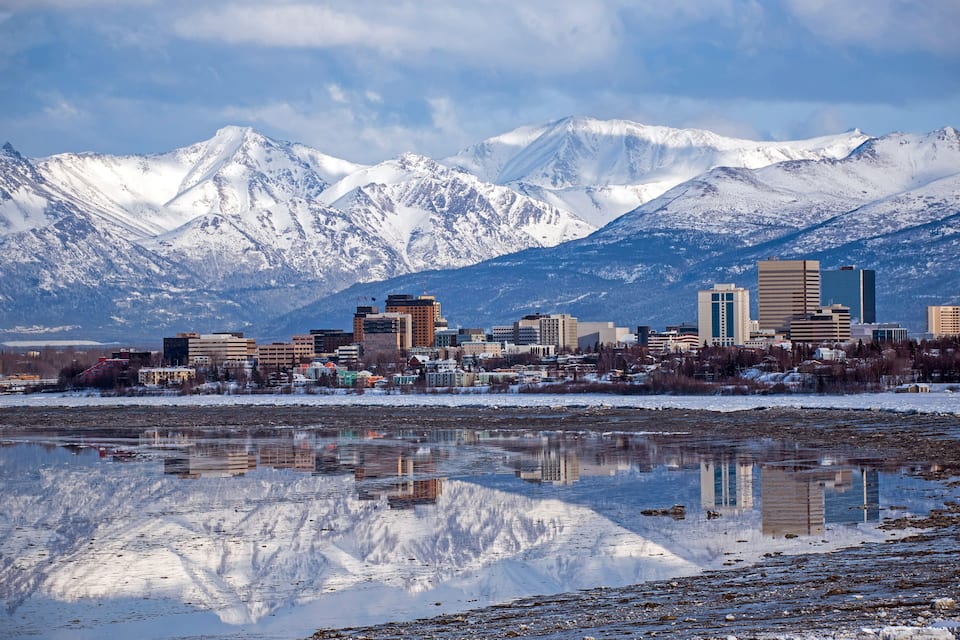 A cityscape with snow-covered mountains in the background and a calm body of water reflecting the buildings and mountains.