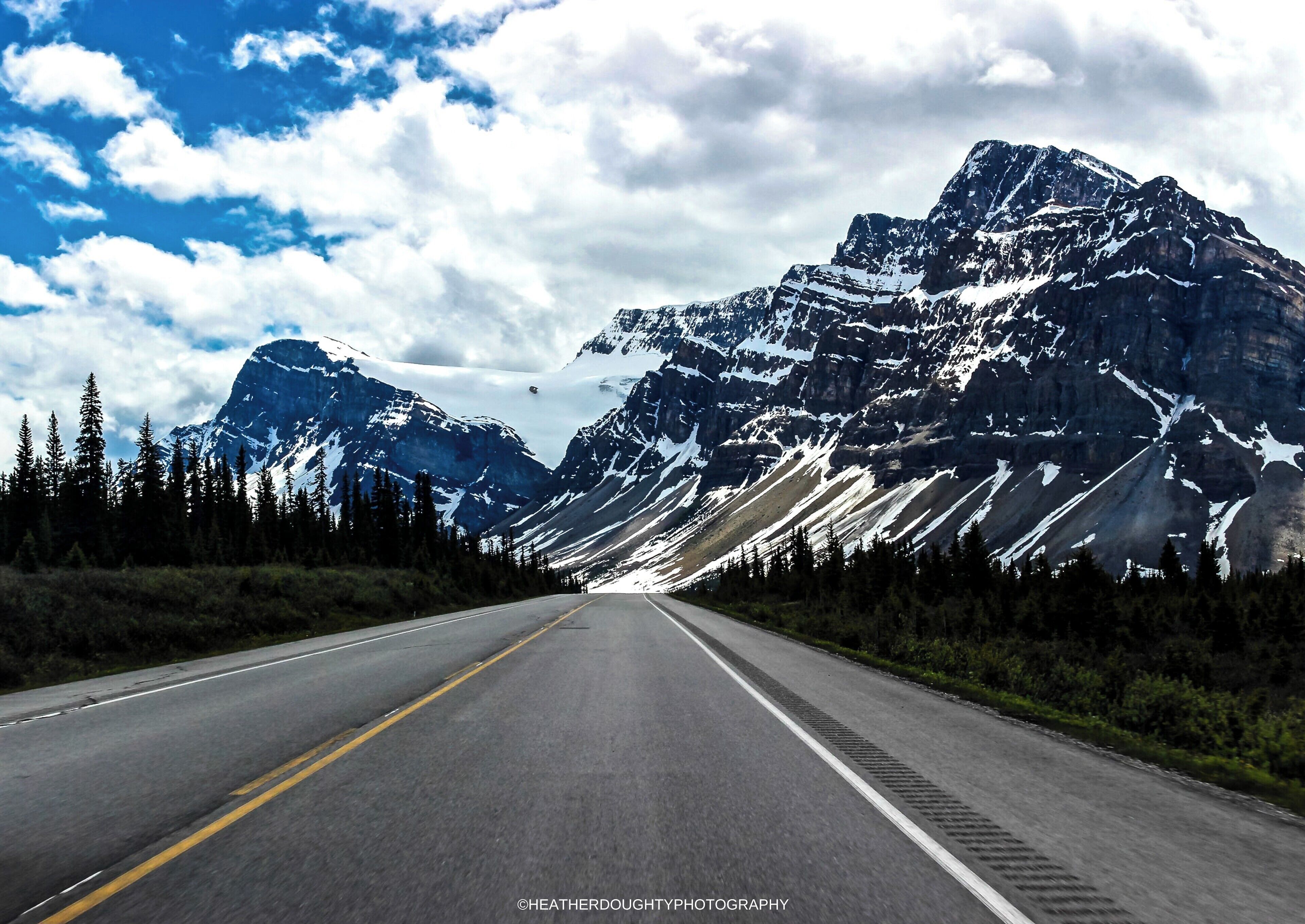 A road flanked by snow-capped mountains and dense forests under a partly cloudy sky.