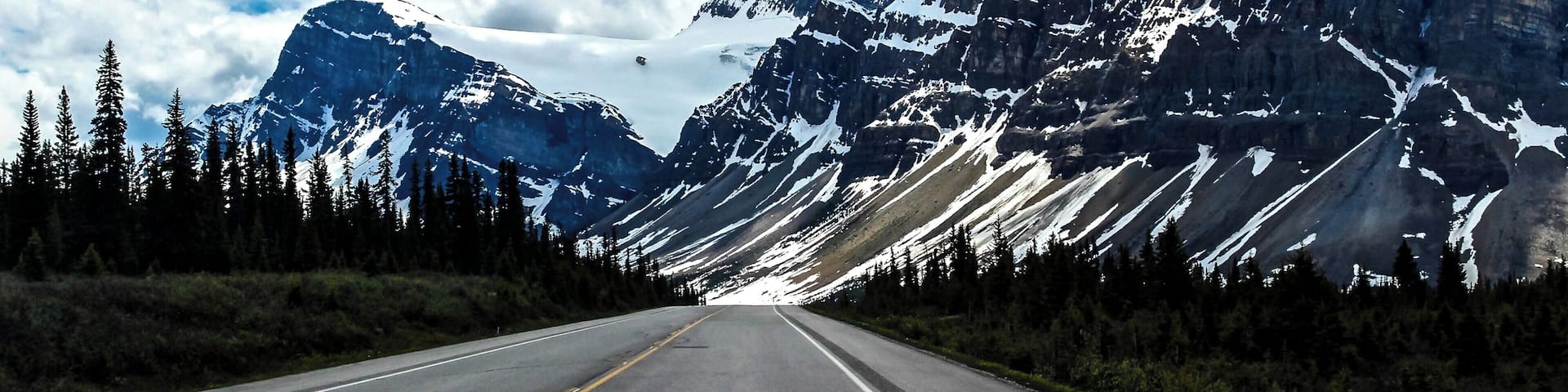 A road flanked by snow-capped mountains and dense forests under a partly cloudy sky.