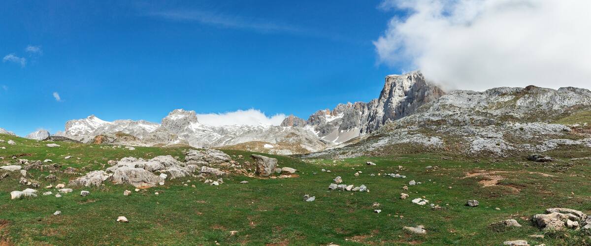 A mountainous landscape with green grass, scattered rocks, and rugged peaks under a clear blue sky.