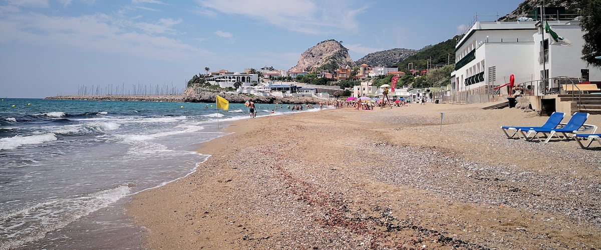 A pebble beach with waves, a rocky hill, and buildings in the background.