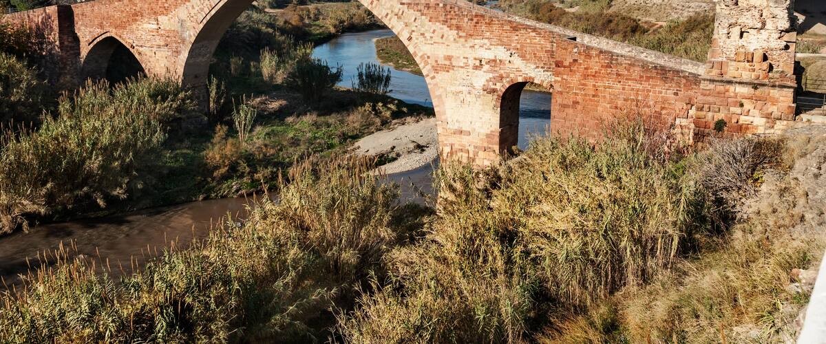 An old stone bridge over a river, surrounded by dry grass and a mountain in the background.