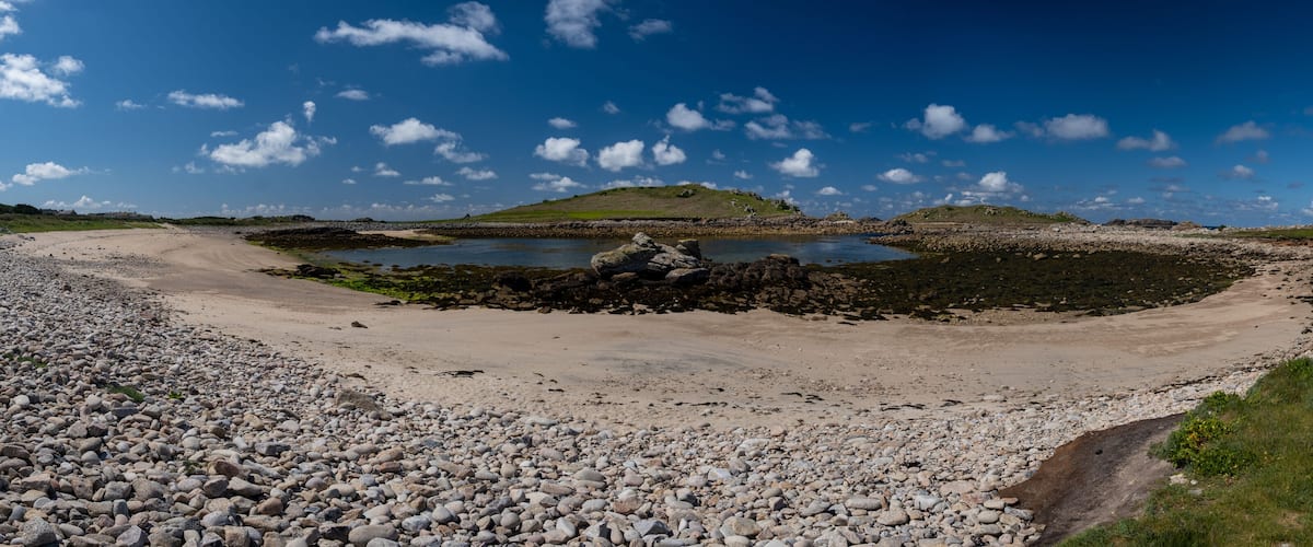 A rocky beach with a small pond surrounded by pebbles and a grassy hill in the background.