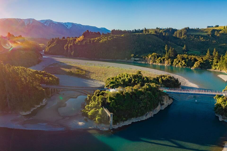 A river winding through a valley with a bridge, surrounded by forested hills and mountains in the background.