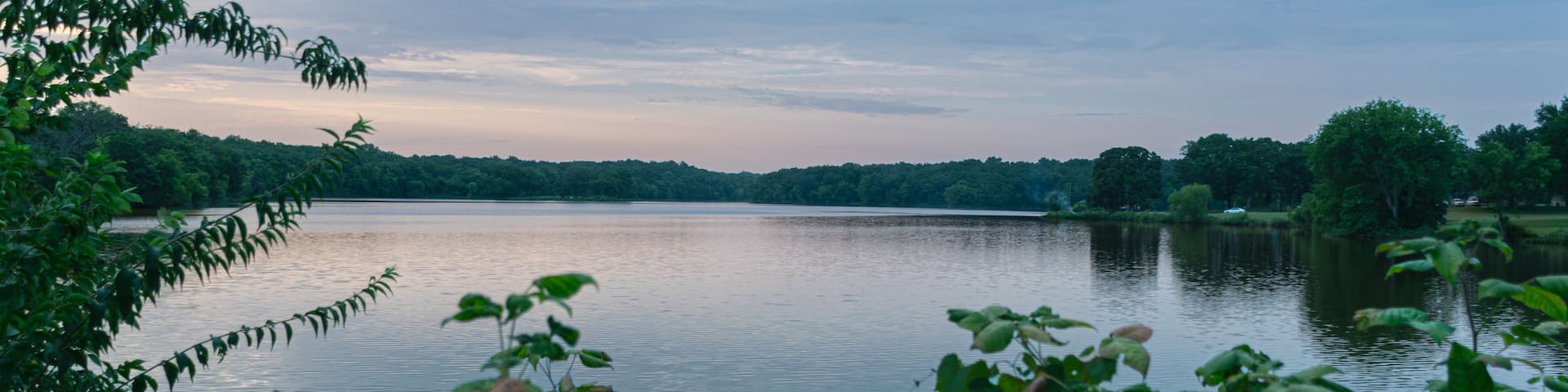 A calm lake surrounded by dense greenery and trees under a cloudy sky.