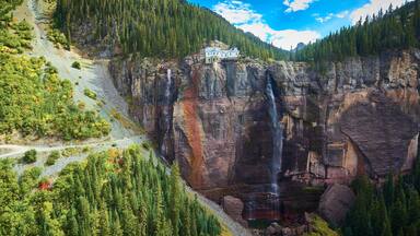 Paisagem montanhosa com uma cachoeira, uma estrada e um edifício em um penhasco.