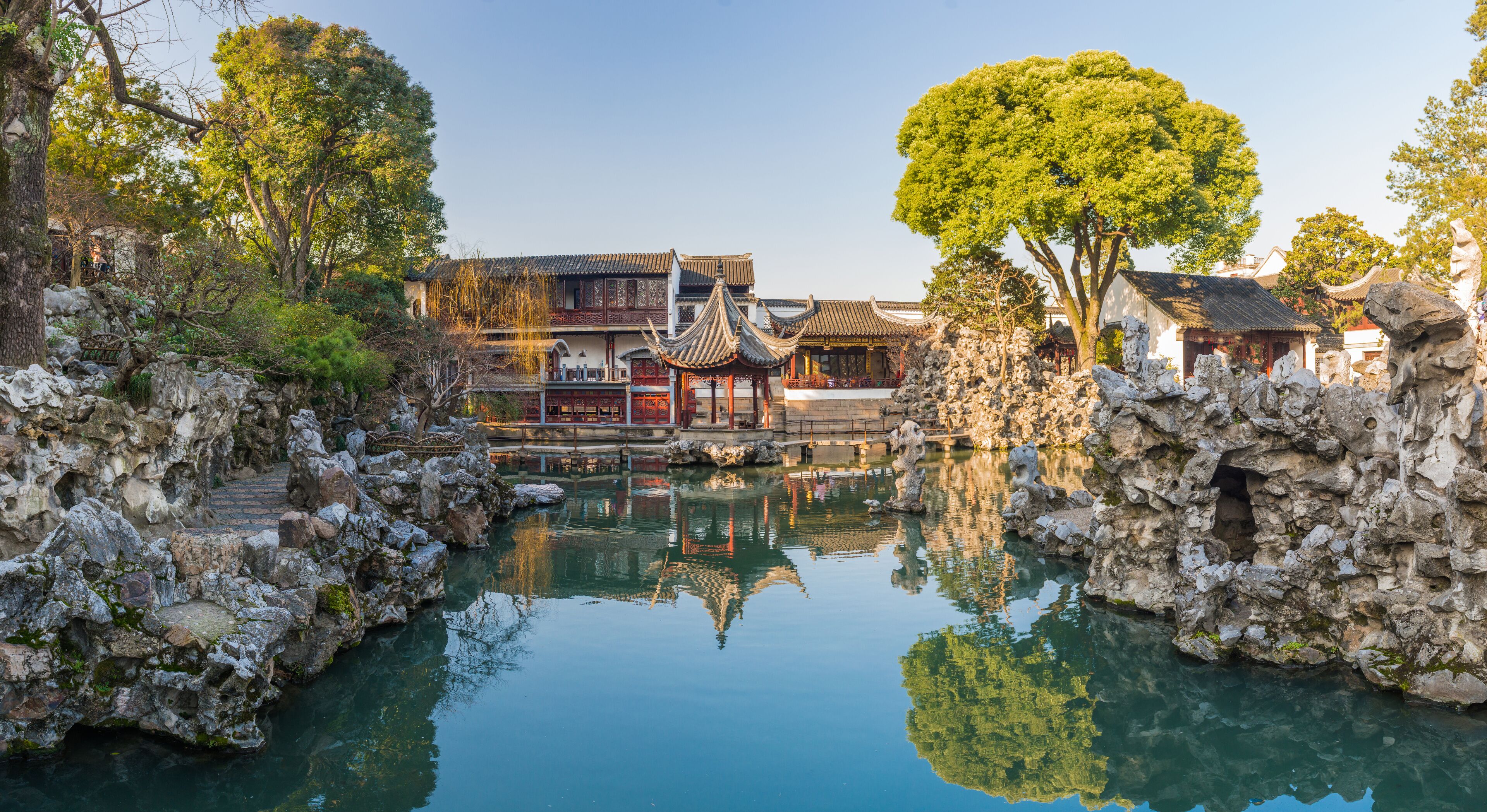 A traditional Chinese courtyard with a clear pond, ornate buildings, and a large tree.
