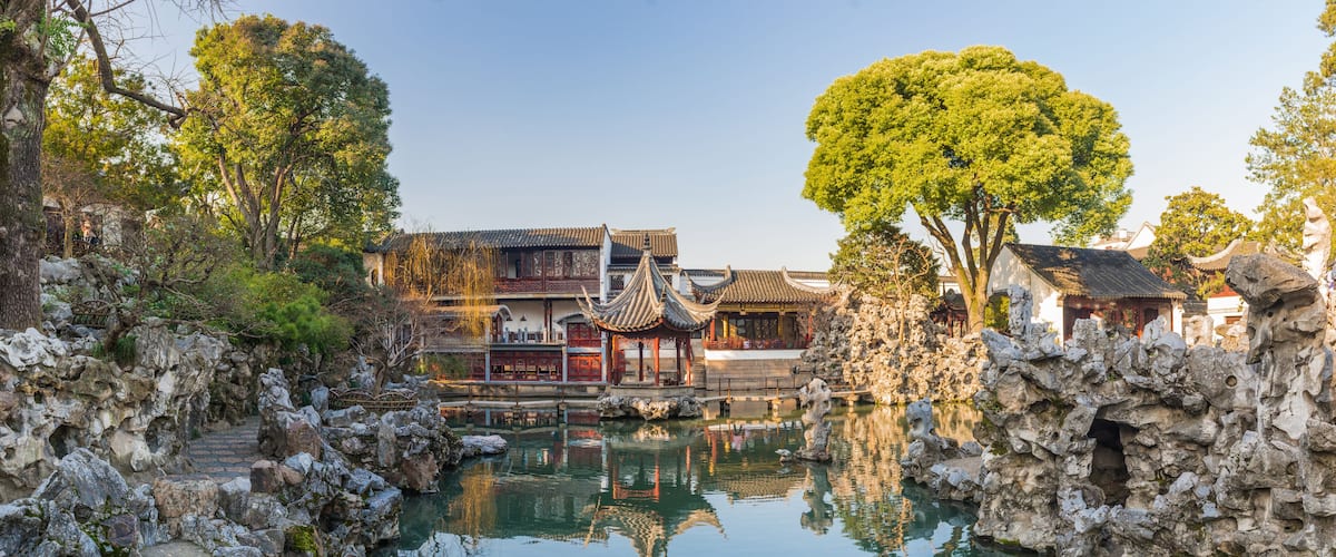 A traditional Chinese courtyard with a clear pond, ornate buildings, and a large tree.