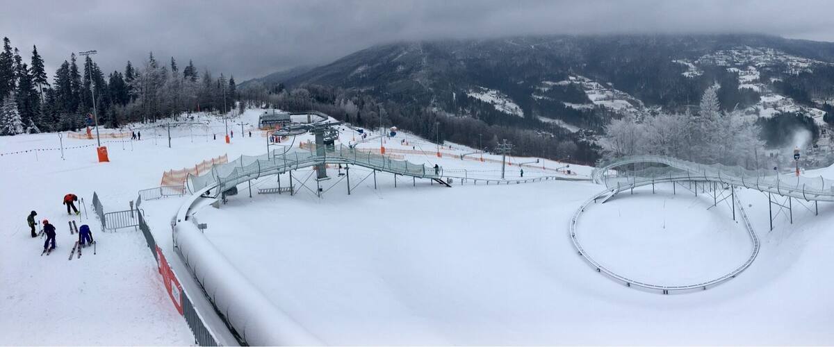 A snow-covered ski slope with a circular track, ski lifts, and a mountainous backdrop.