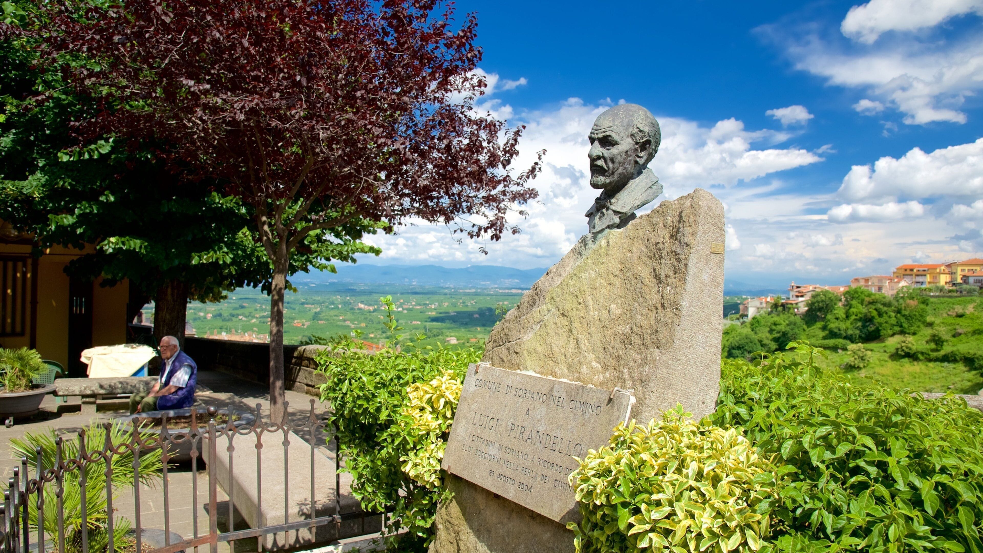 A statue of Luigi Pirandello, surrounded by greenery and a scenic view of a town in the background.