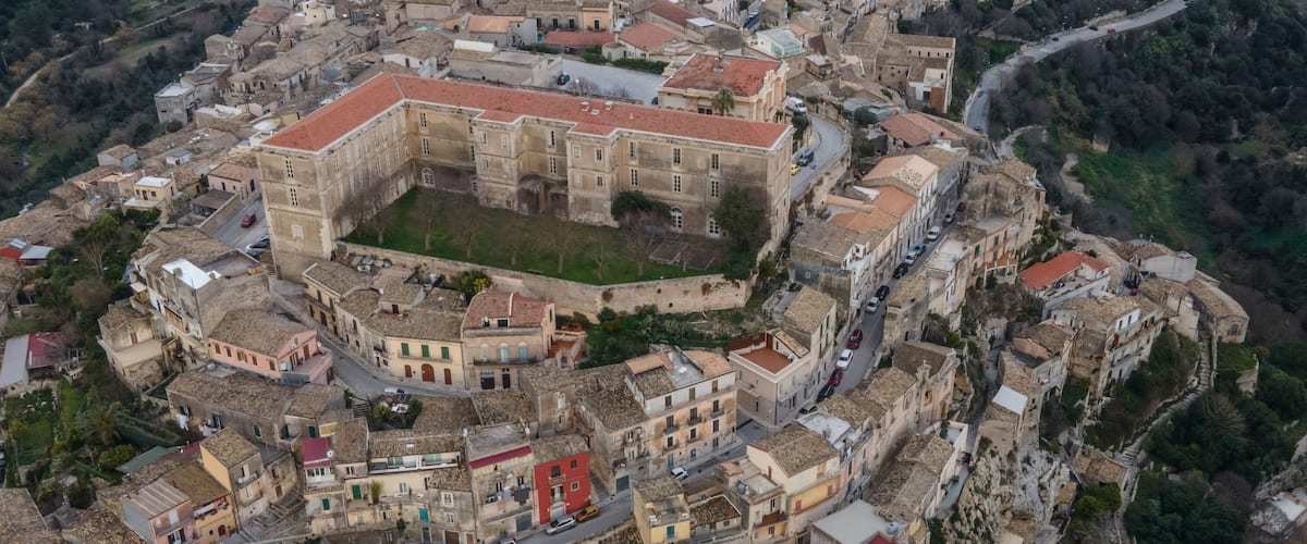 An aerial view of a historic town with a prominent central fortress, winding roads, and surrounding greenery.