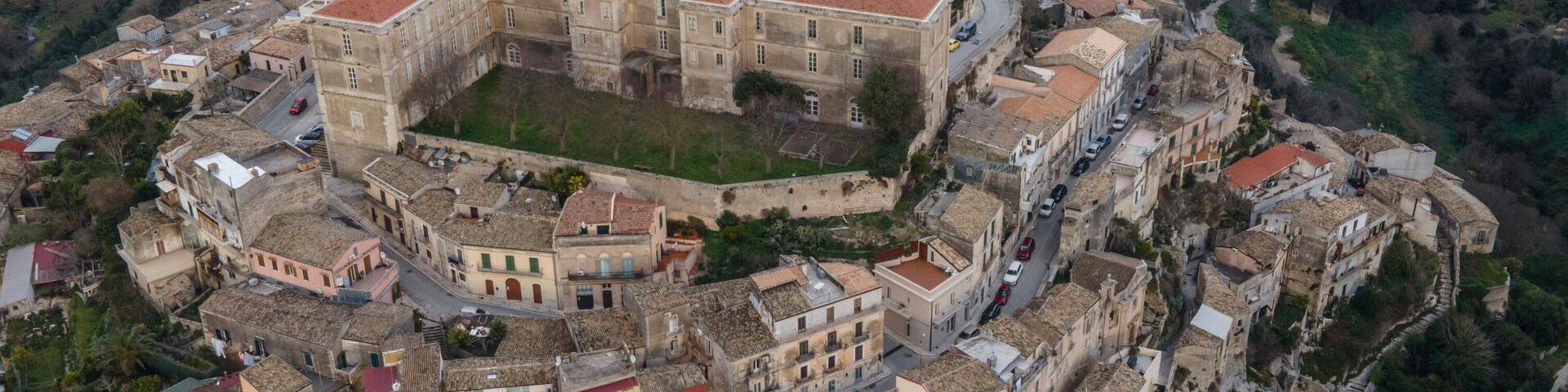 An aerial view of a historic town with a prominent central fortress, winding roads, and surrounding greenery.