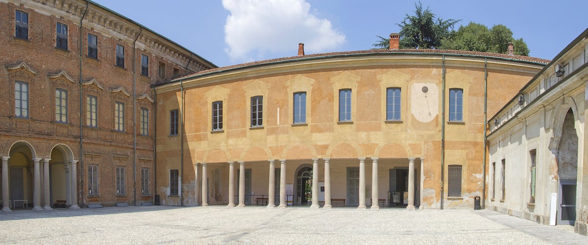 A courtyard with a yellow building featuring columns and arched doorways, surrounded by a cobblestone ground.