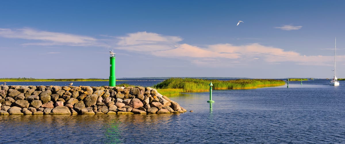 A green lighthouse, a rocky breakwater, and a sailboat on a calm water body with distant greenery and a clear sky.