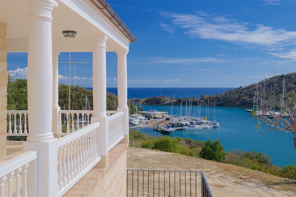 Vista costera con un edificio blanco, un puerto deportivo con barcos y un paisaje montañoso.