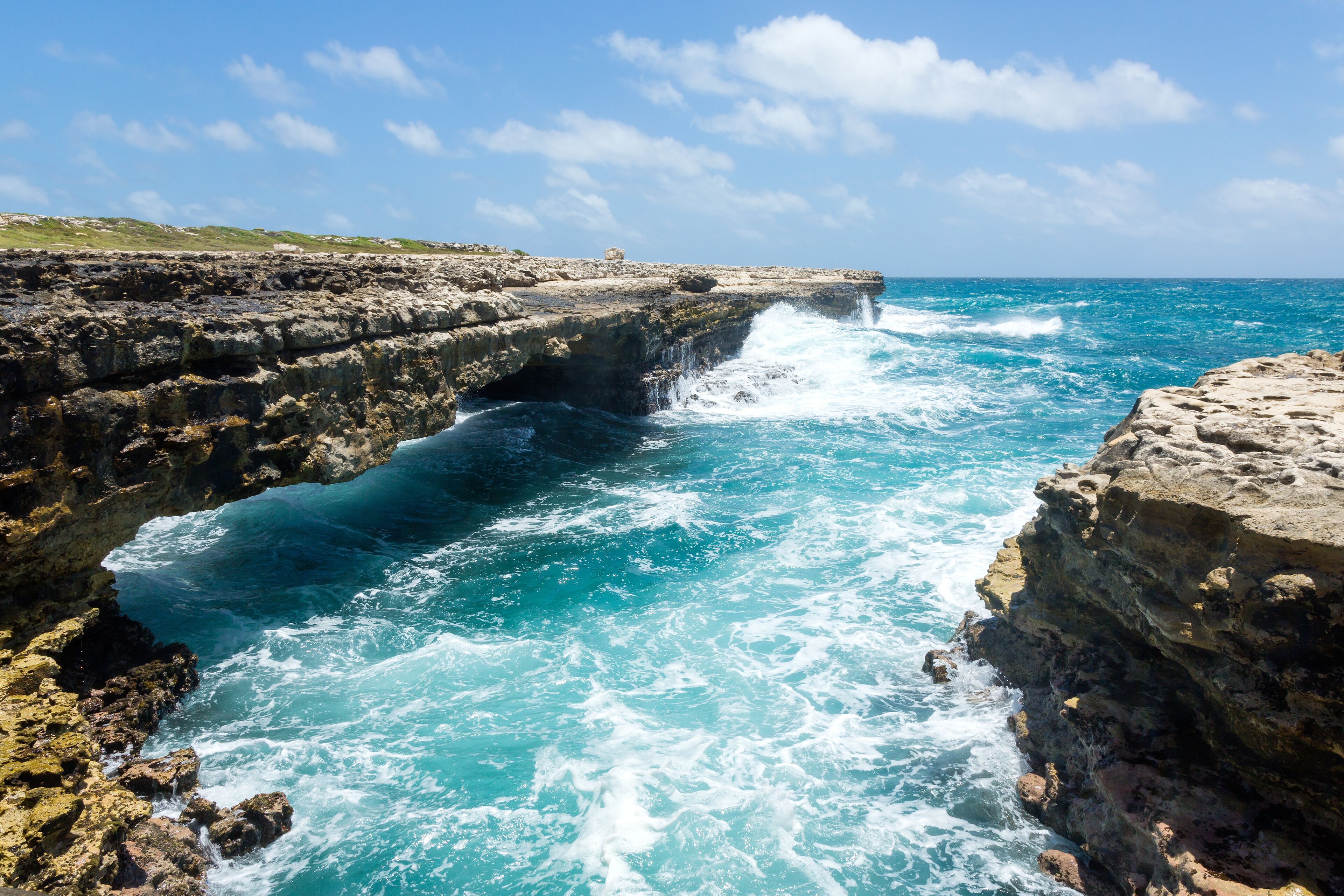 A rocky coastline with waves crashing against the rocks, a clear blue sky, and a distant view of the horizon.