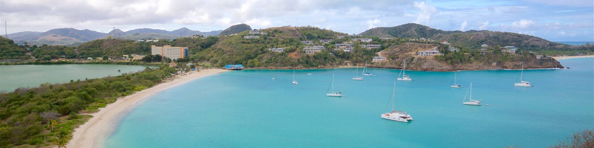 A coastal bay with turquoise waters, several sailboats, a sandy beach, and buildings on hills in the background.