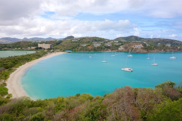 Une baie côtière aux eaux turquoise, avec plusieurs voiliers, une plage de sable et des bâtiments situés sur des collines en arrière-plan.