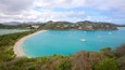 Una bahía costera con aguas turquesas, varios veleros, una playa de arena y edificios en las colinas al fondo.