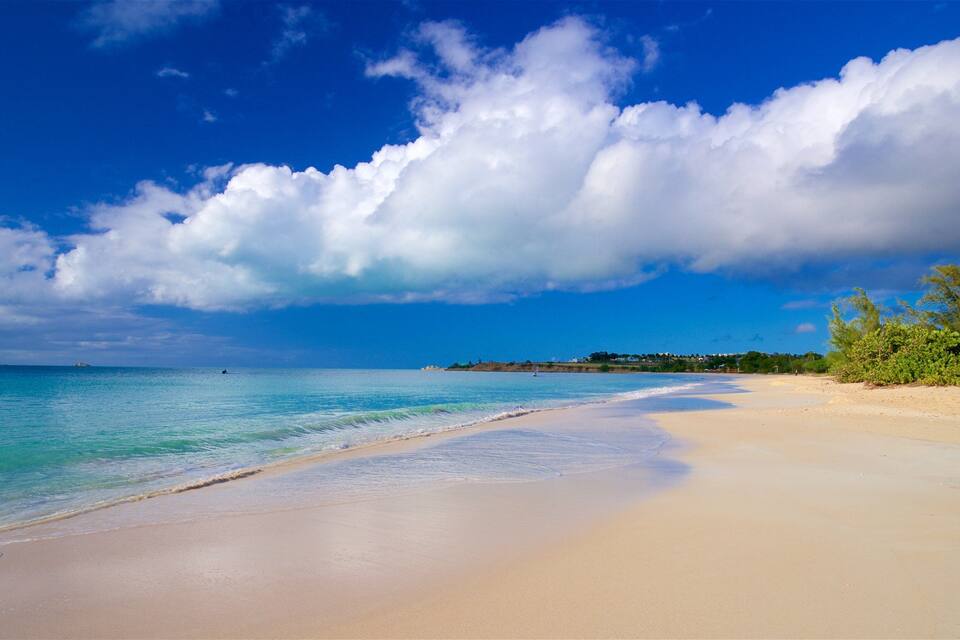 A beach with clear turquoise water, a sandy shore, and a distant shoreline with buildings.