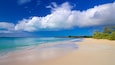 Ein Strand mit klarem türkisblauem Wasser, einem sandigen Ufer und einer fernen Küstenlinie mit Gebäuden.