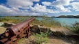 Eine verrostete Kanone auf felsigem Ufer mit Blick auf einen türkisblauen See, im Hintergrund Berge.