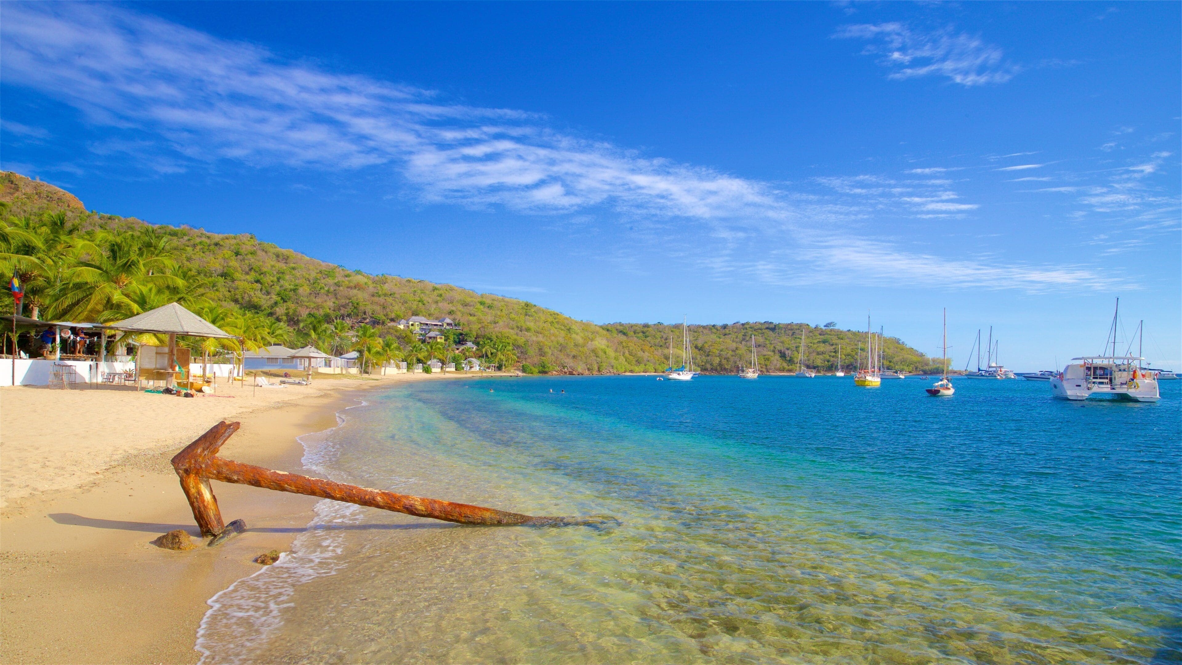 A beach with clear turquoise waters, a large rusted anchor on the shore, and several boats anchored in the distance.