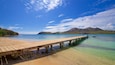 Un muelle de madera que se adentra en un mar turquesa transparente, con una playa de arena y montañas a lo lejos.