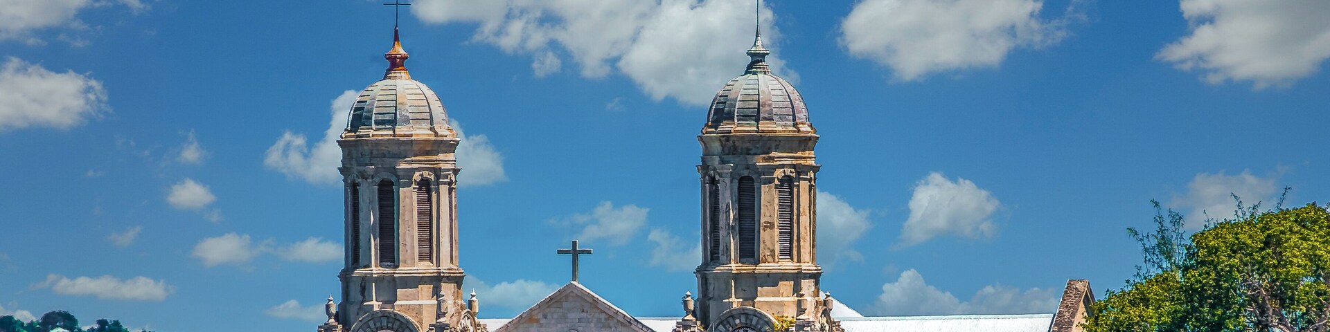 Una iglesia histórica con dos torres abovedadas y una torre del reloj, rodeada de abundante vegetación.