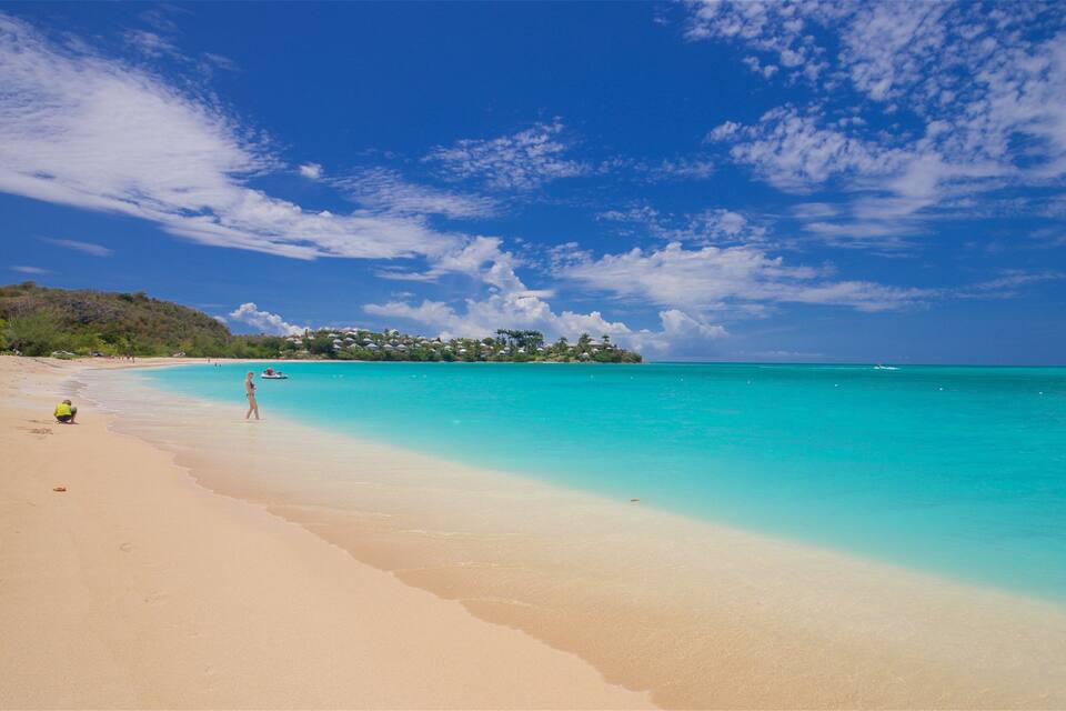 A beach with clear turquoise waters, a sandy shore, and a small island with buildings in the background.