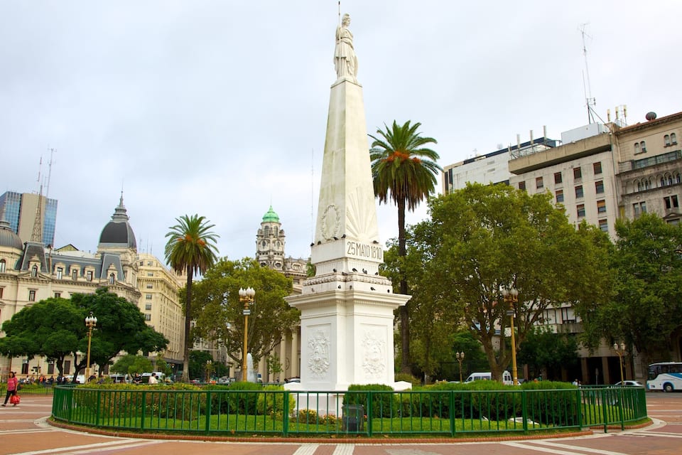 A monument with a date inscription, surrounded by greenery and urban buildings.