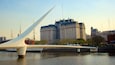 Un puente moderno con un arco blanco sobre un río, con edificios históricos al fondo.