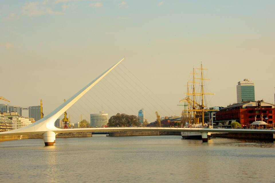 Eine moderne Brücke mit weißem Bogen über einem Fluss, umgeben von Stadtgebäuden und einem nahegelegenen Schiff.