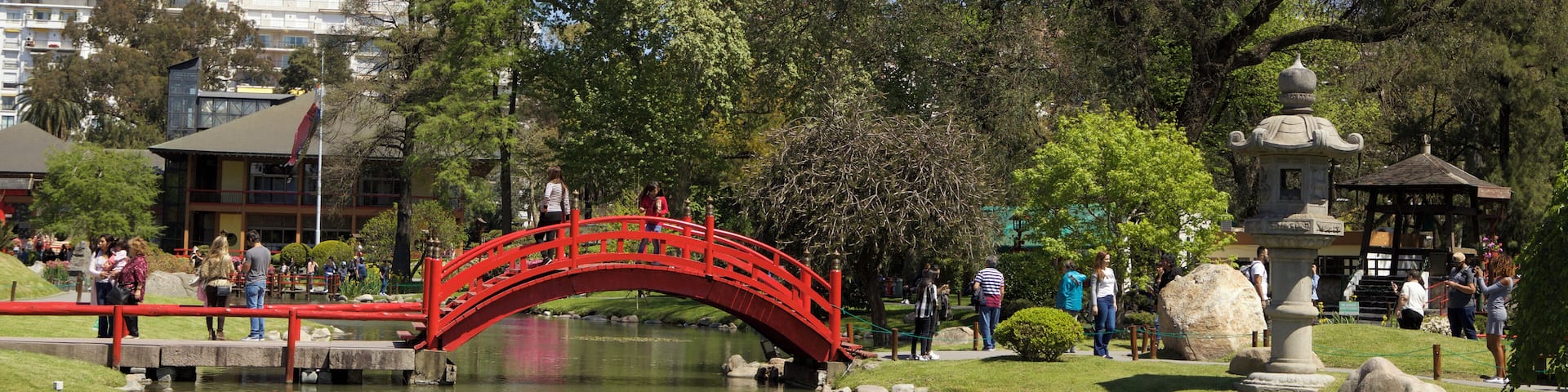 Un puente rojo sobre un estanque cubierto de lirios, rodeado de árboles y una pagoda de piedra.