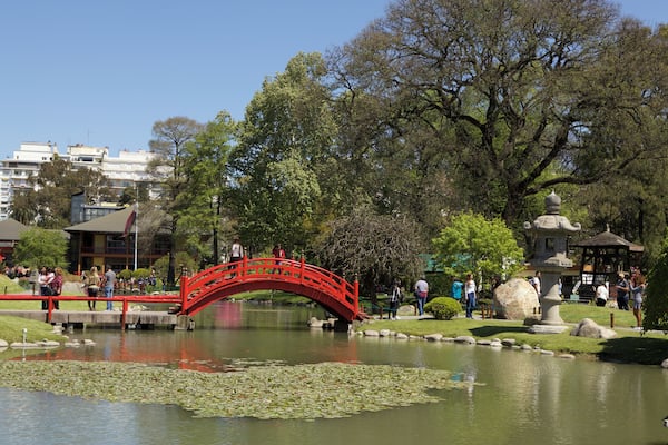 Eine rote Brücke über einen Teich mit Seerosen, umgeben von Bäumen und einer steinernen Pagode.