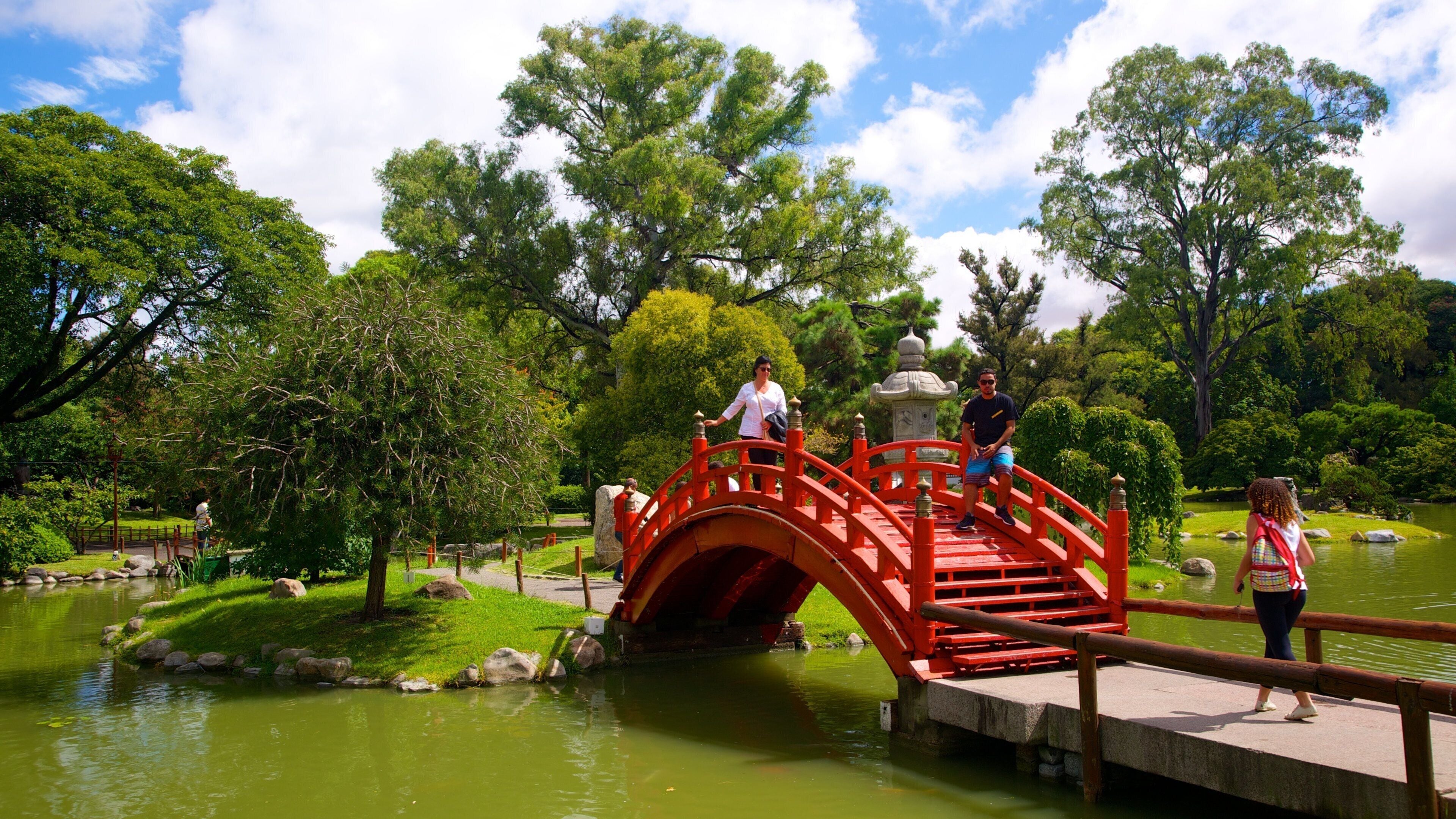 Un puente rojo sobre un estanque en un parque con abundante vegetación y árboles.