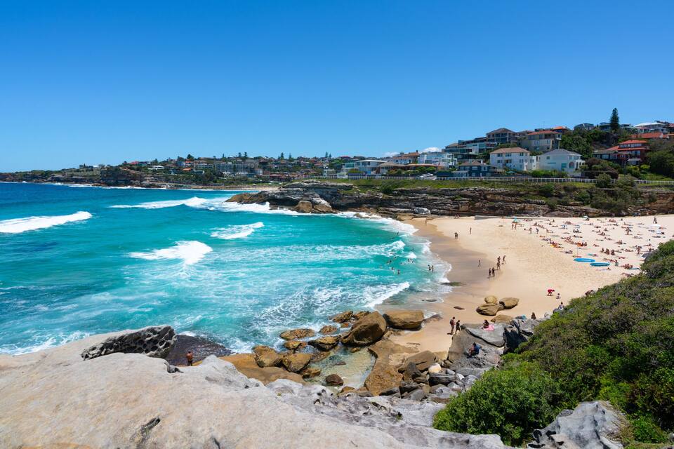 A coastal beach with waves, a sandy shore, and buildings in the background.