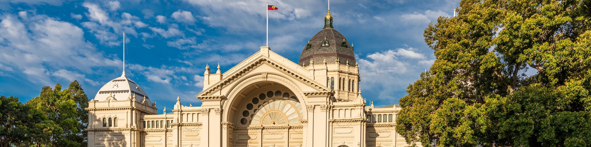 Un edificio grande y ornamentado con una cúpula destacada y dos mástiles con banderas, rodeado de abundante follaje.