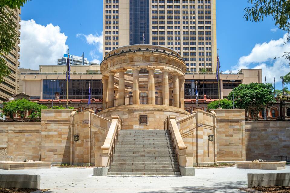 A grand entrance with a circular colonnade, a staircase leading up to it, and a tall building in the background.