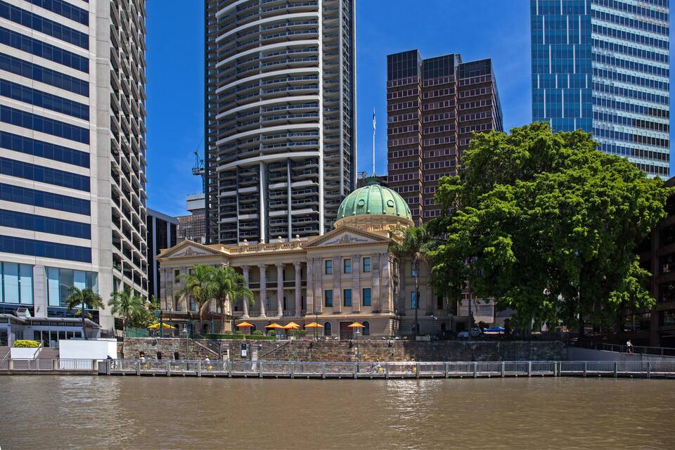 A waterfront cityscape with a prominent building featuring a green dome, surrounded by modern skyscrapers and palm trees.