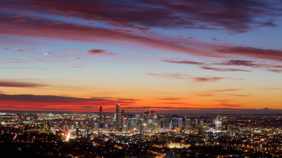 A cityscape at dusk with a colorful sky, illuminated buildings, and a crescent moon.