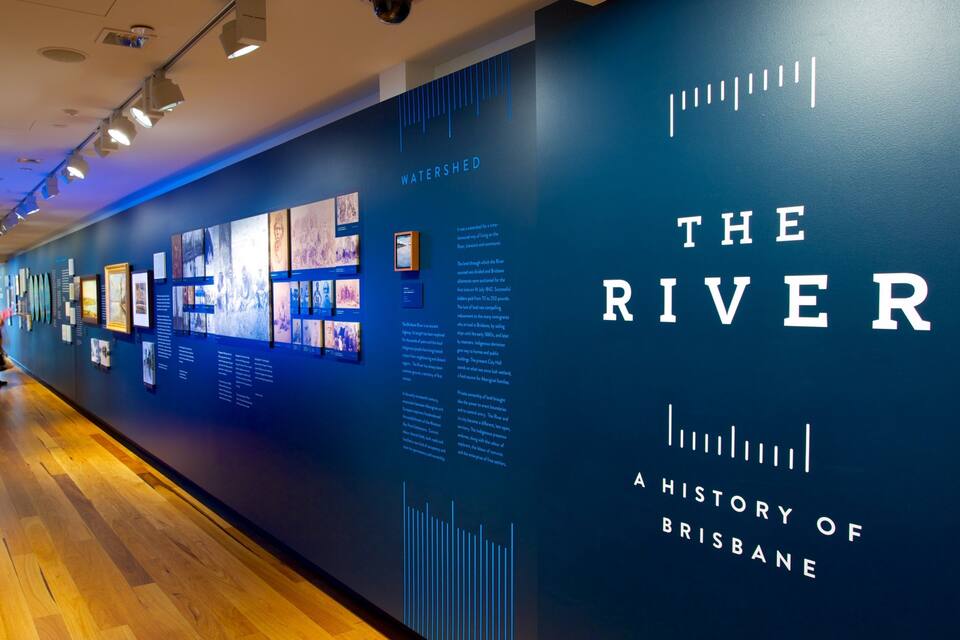An indoor exhibition with a large blue wall displaying 'The River: A History of Brisbane' and various photographs and information panels.