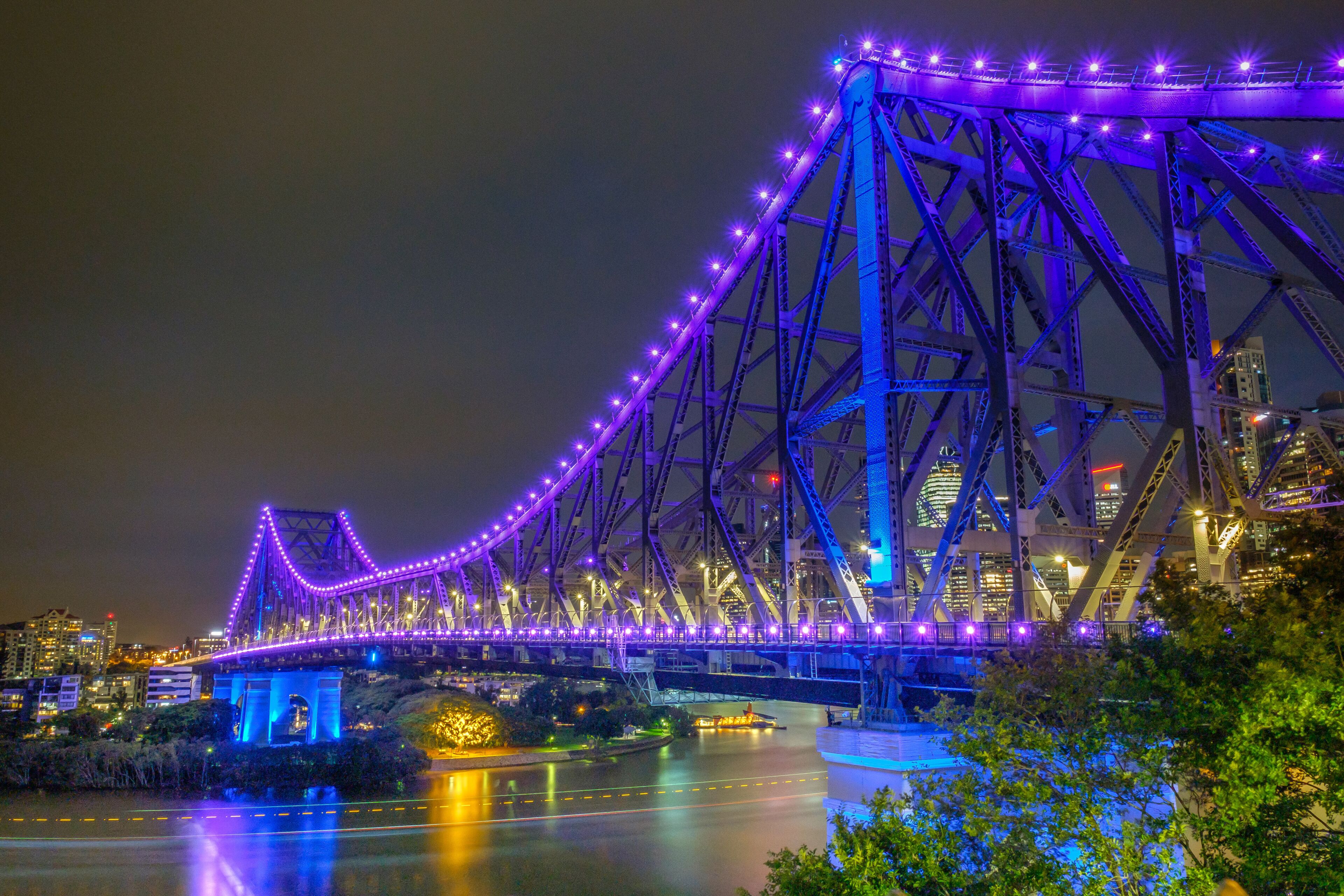 A bridge with purple lights at night, surrounded by city buildings and a river.